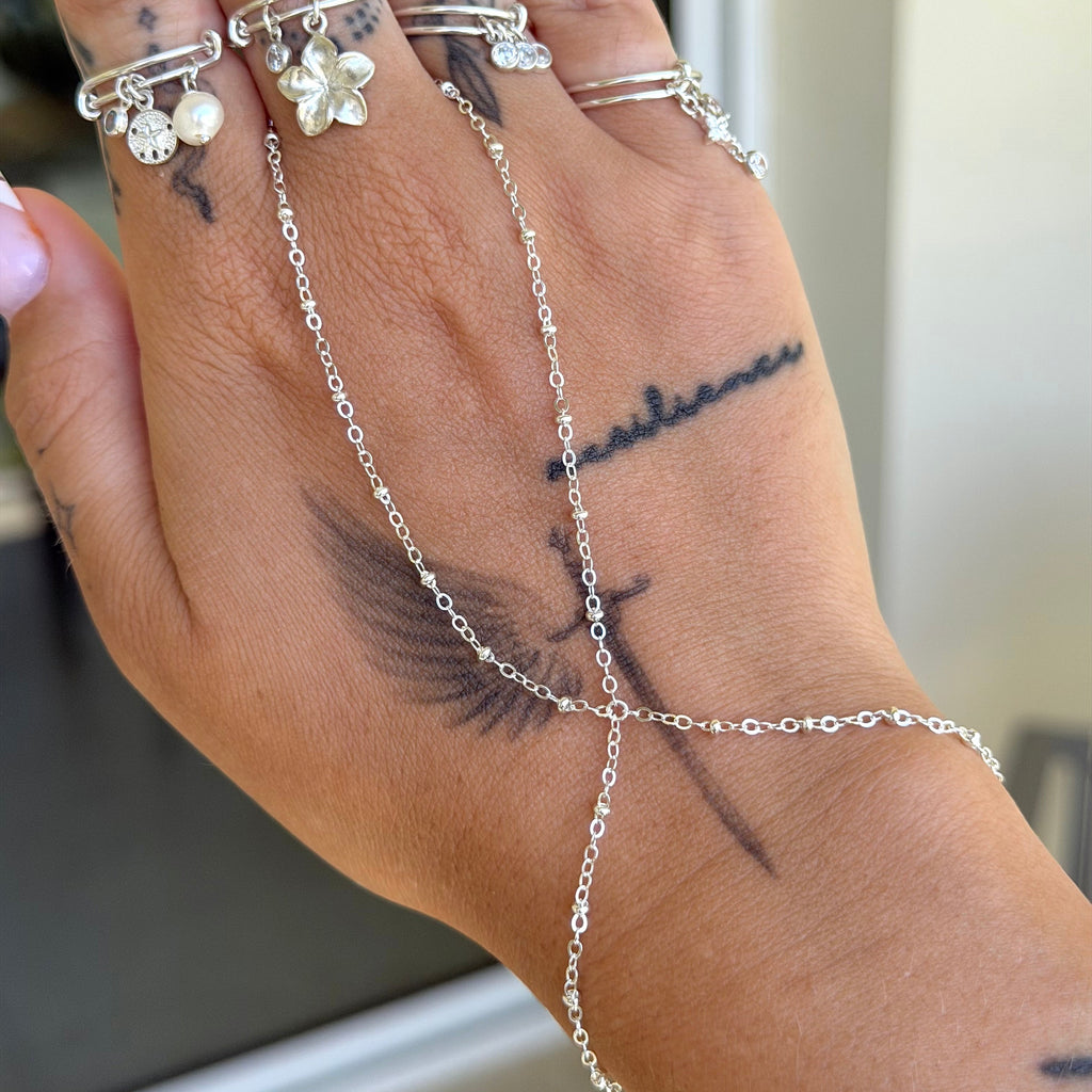 A close-up of a hand wearing multiple silver rings and a matching silver chain hand bracelet/anklet set, with visible tattoos on the skin.