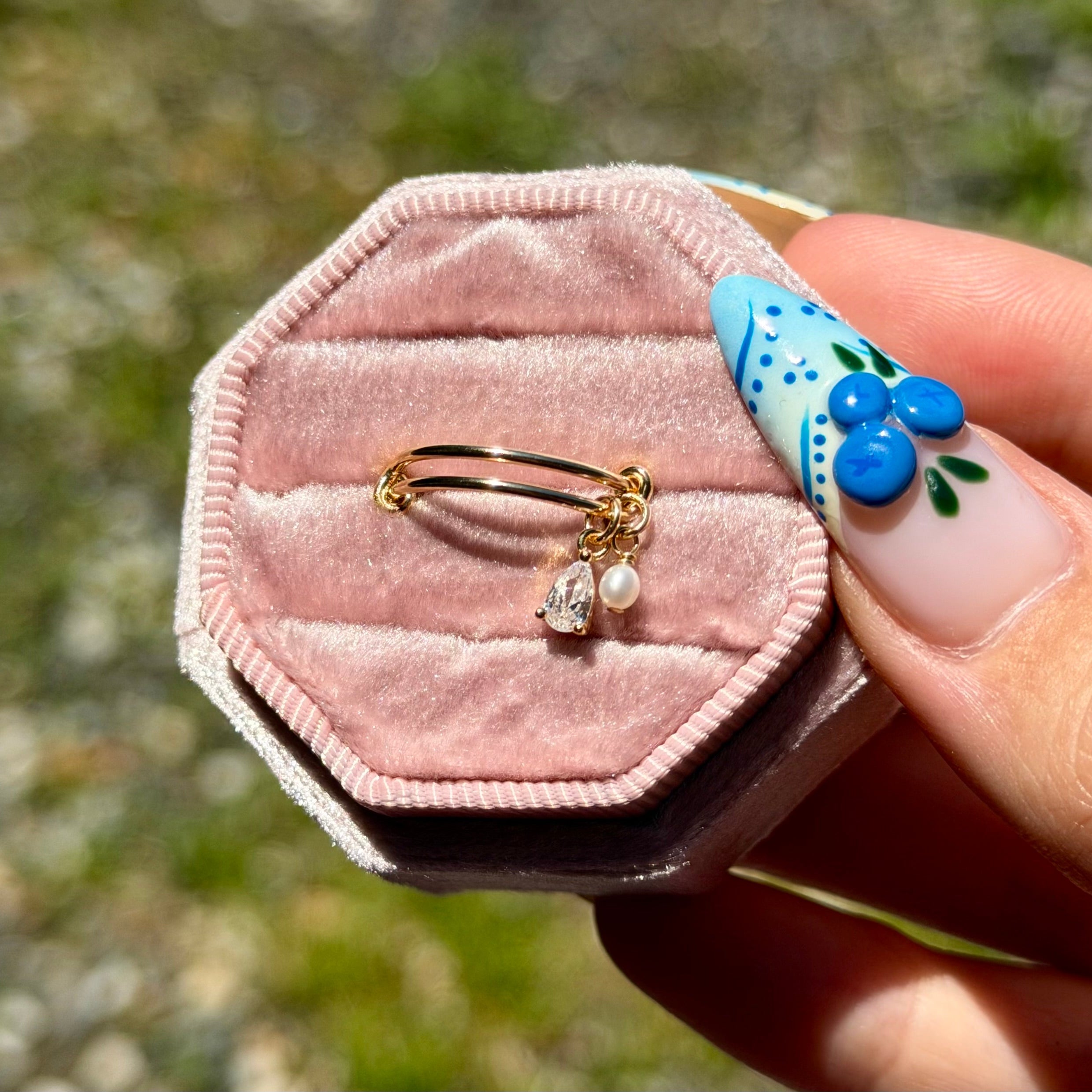 Pink jewelry box with gold ring and pearl pendant held by a hand with blue nail polish.