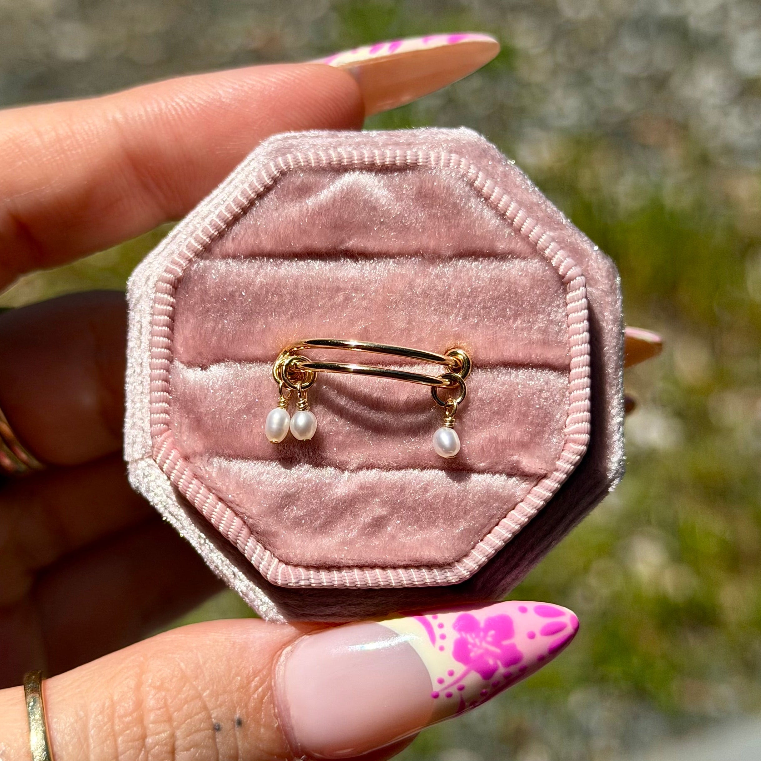 Pink velvet jewelry box with a gold ring and pearls held by a hand with floral nail polish.