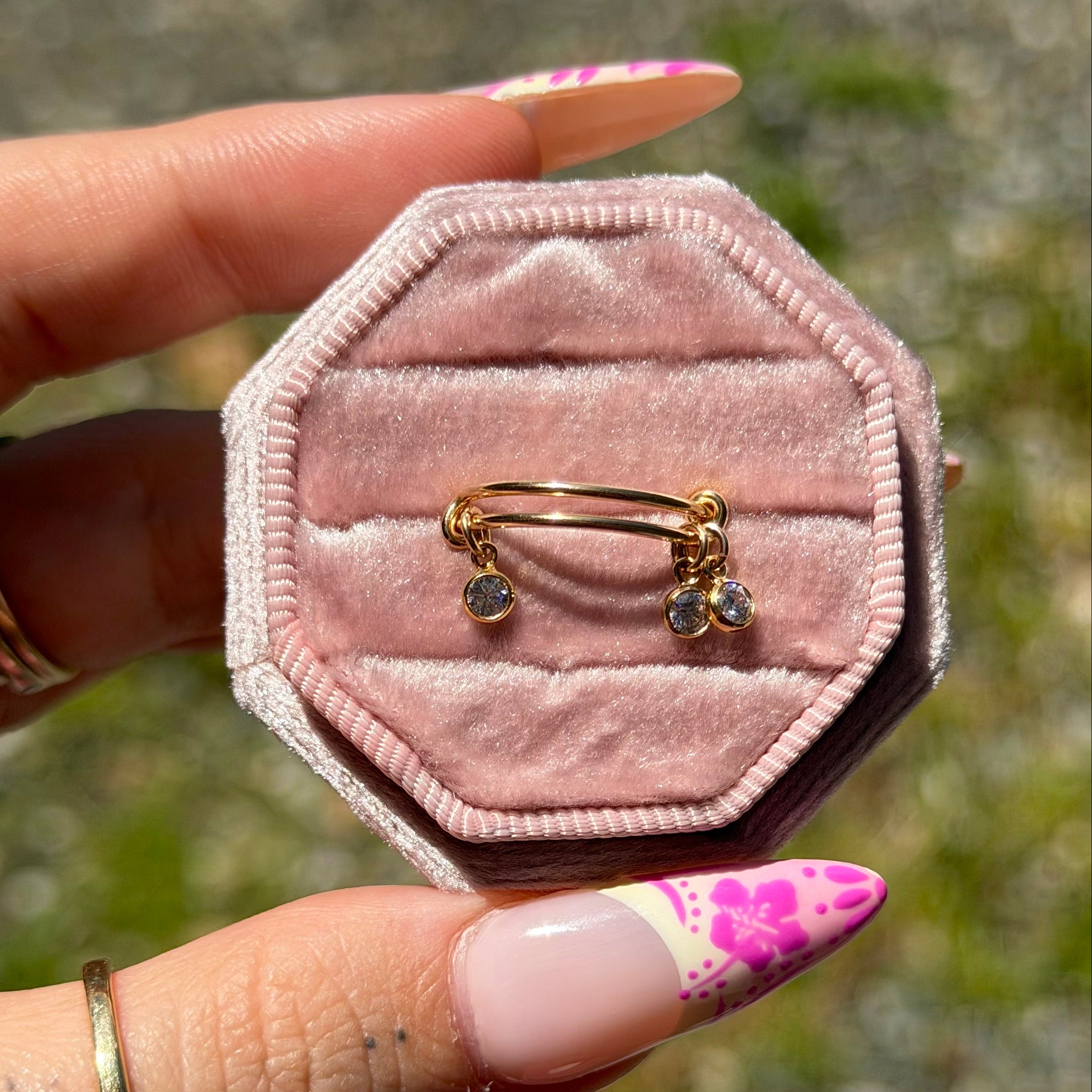 Pink velvet jewelry box with gold ring inside, held by a hand with floral nail polish.