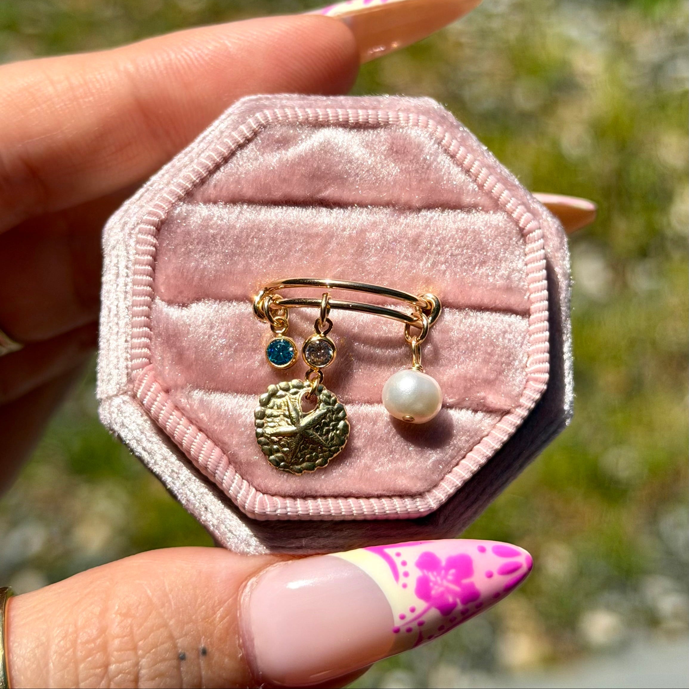 Pink hexagonal jewelry box with gold ring and charms held by a hand with pink nail polish.