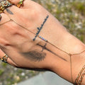 Hand with multiple gold rings and bracelets, featuring tattoos on the palm and fingers, against a blurred natural background.