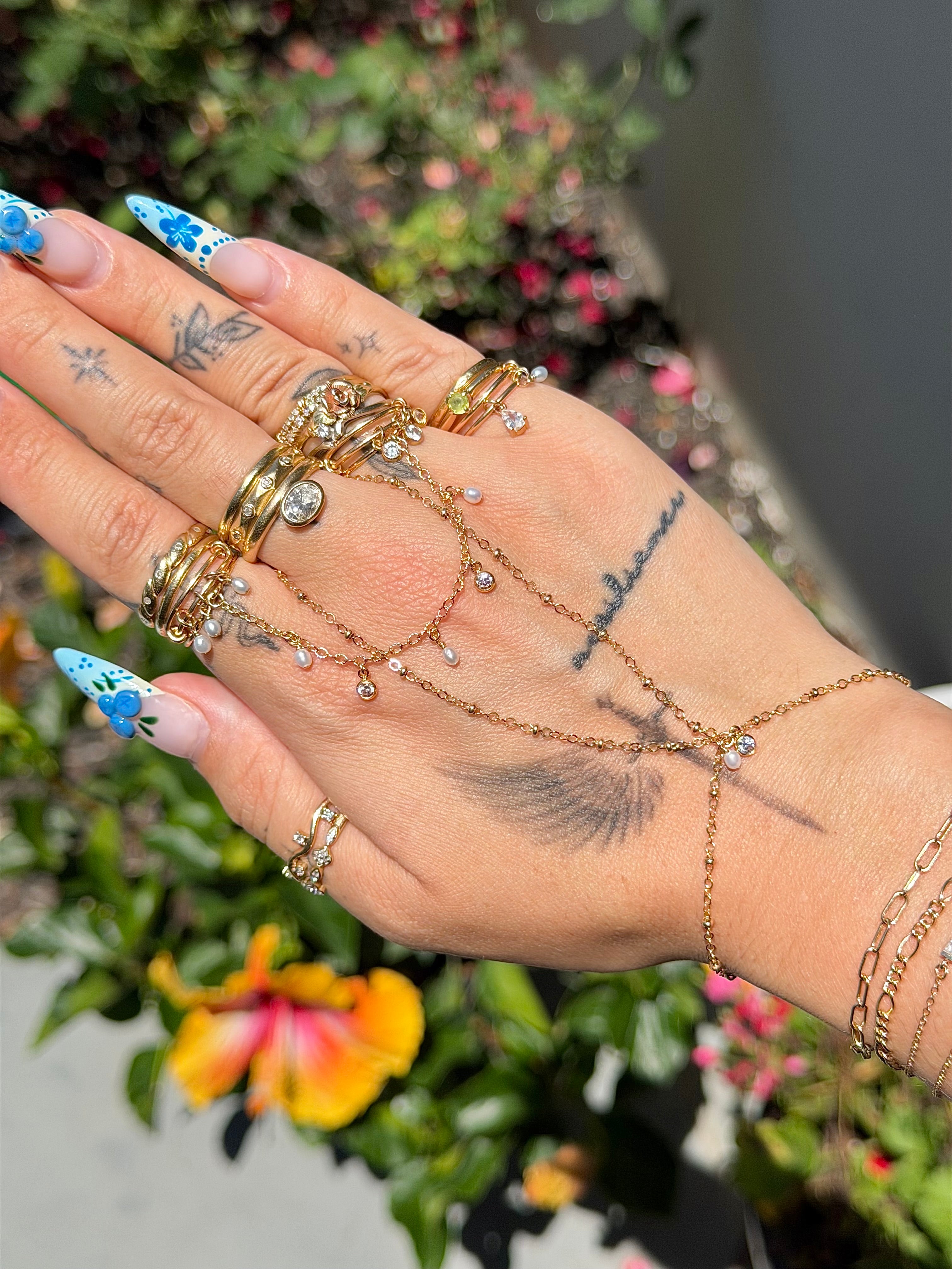 Hand wearing multiple gold rings and bracelets with floral nail designs against a blurred natural background