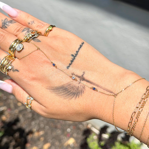 Hand with multiple gold rings and bracelets, featuring tattoos, against a blurred outdoor background.
