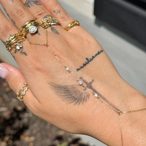 Hand with multiple gold rings and a delicate gold chain, set against a blurred outdoor background.