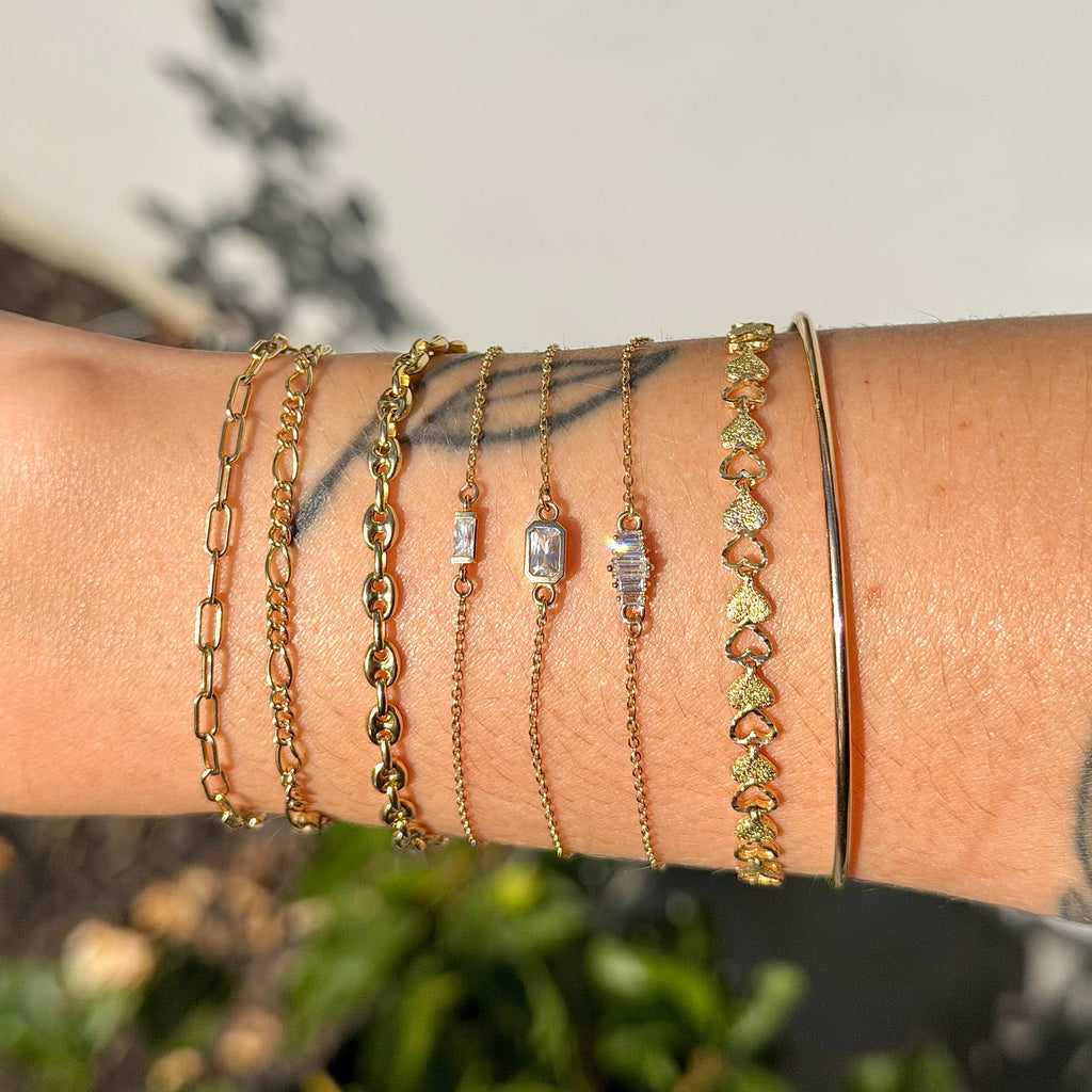 Close-up of a wrist wearing multiple gold bracelets with green gemstones against a blurred natural background.