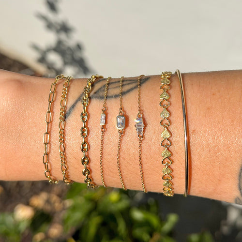 Close-up of a wrist wearing multiple gold bracelets with green gemstones against a blurred natural background.